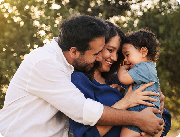 Família de três pessoas se abraçando ao ar livre, sorrindo; criança de camisa azul.
