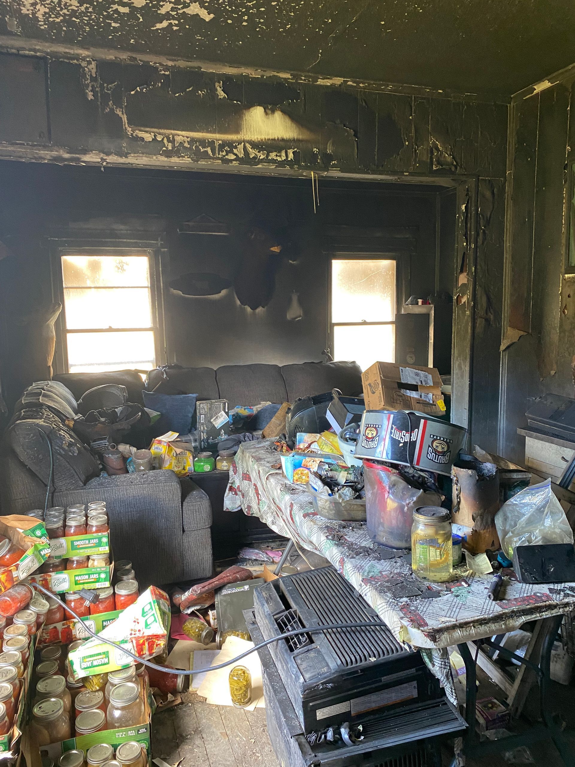 Fire-damaged room with charred walls, debris, and stacked supplies in front of two windows.