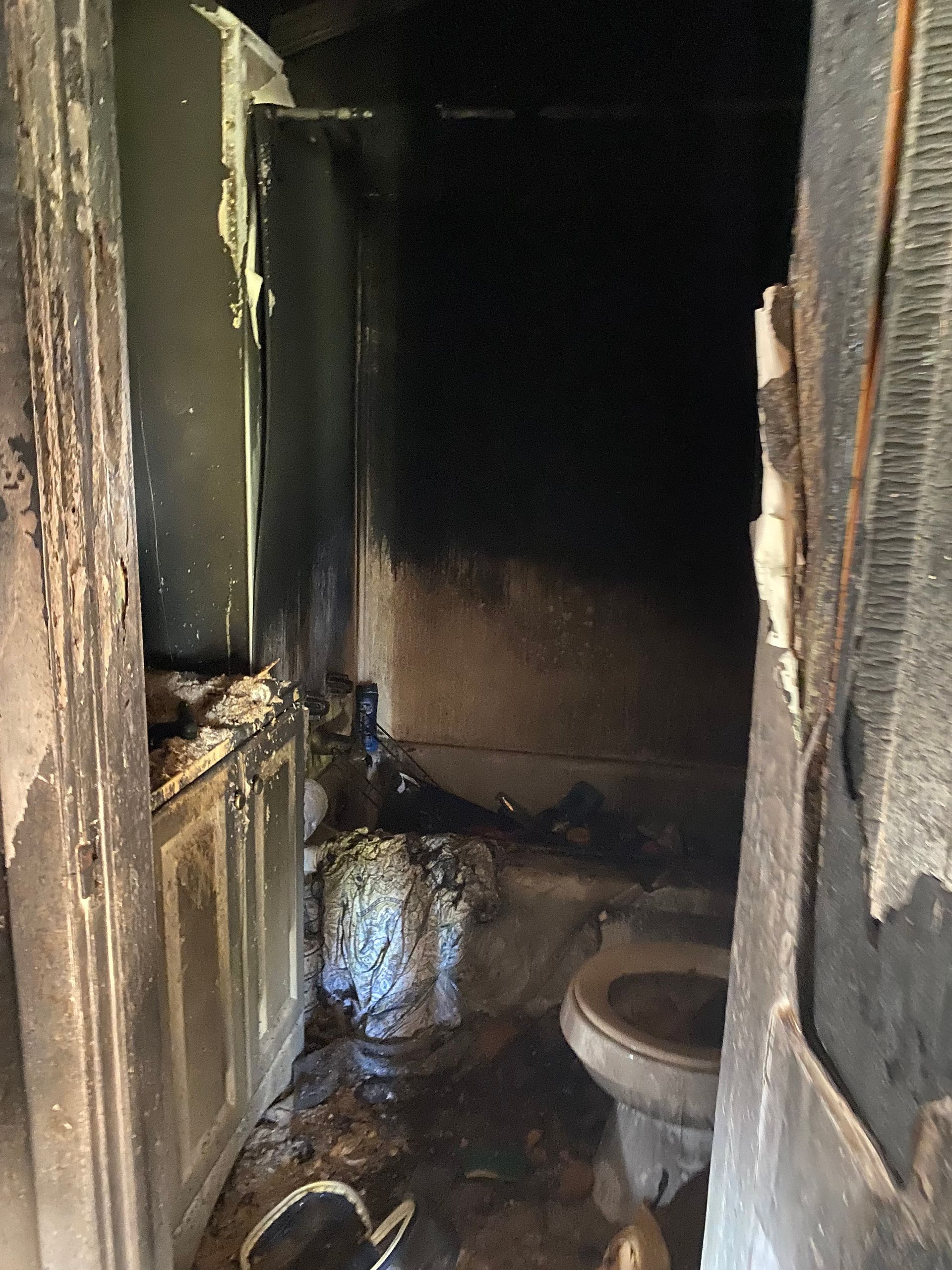 Charred narrow hallway leading to a small bathroom with a toilet, showing fire damage and soot-covered walls