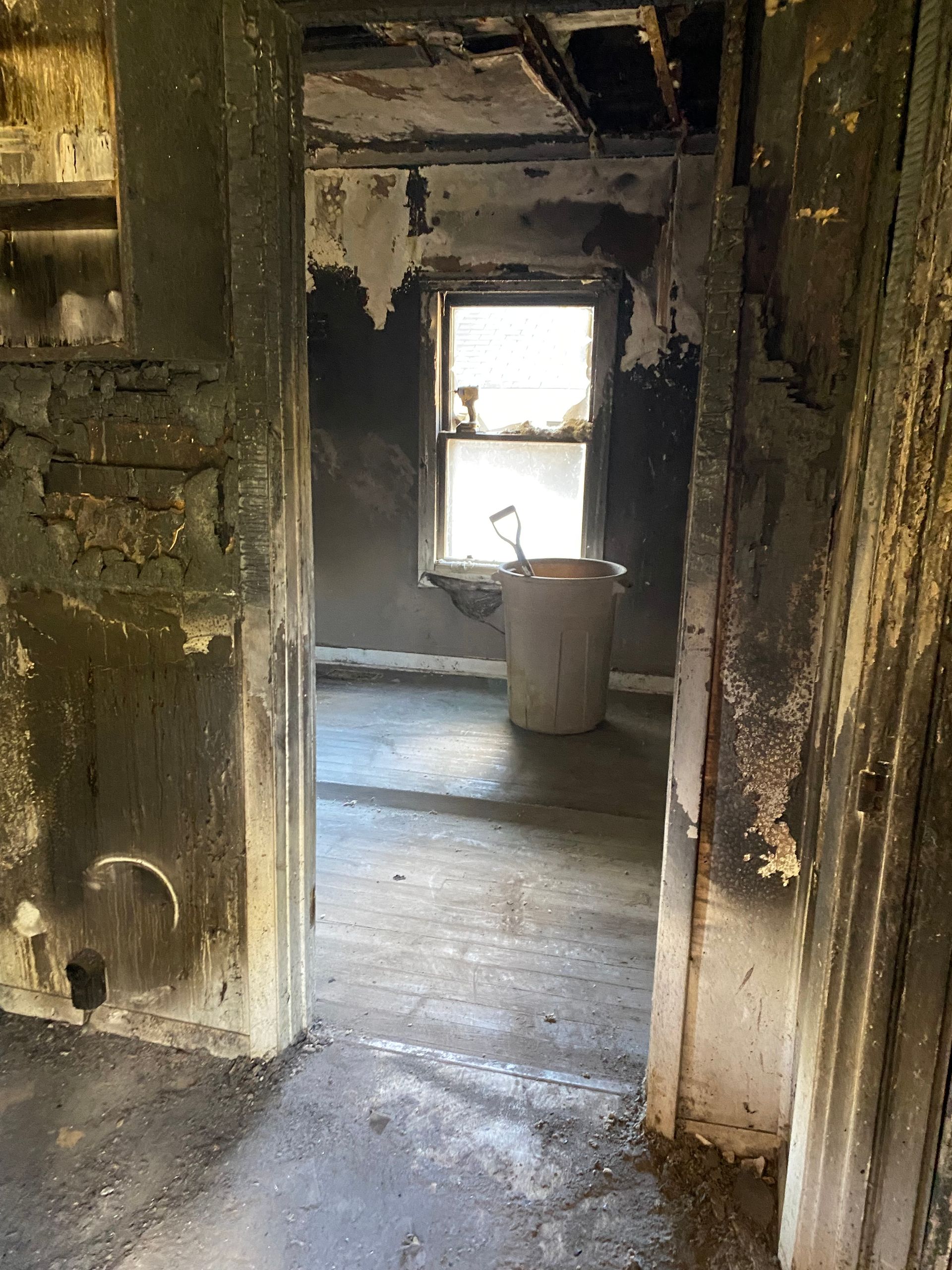 Charred, damaged interior room with peeling walls, wet floor, and a bucket near a bright window.