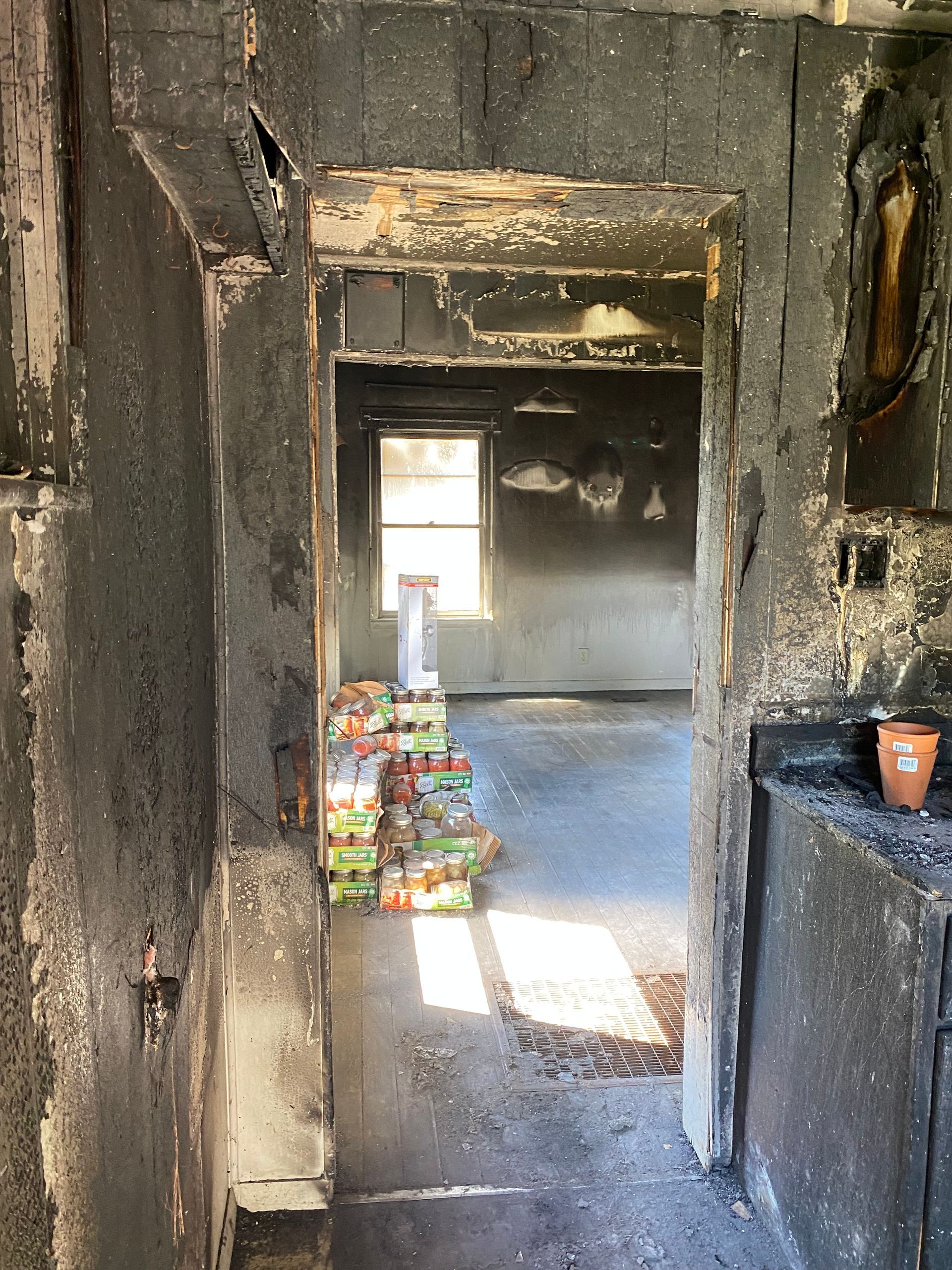 Charred doorway leading to a dim room with stacked groceries and a small potted plant