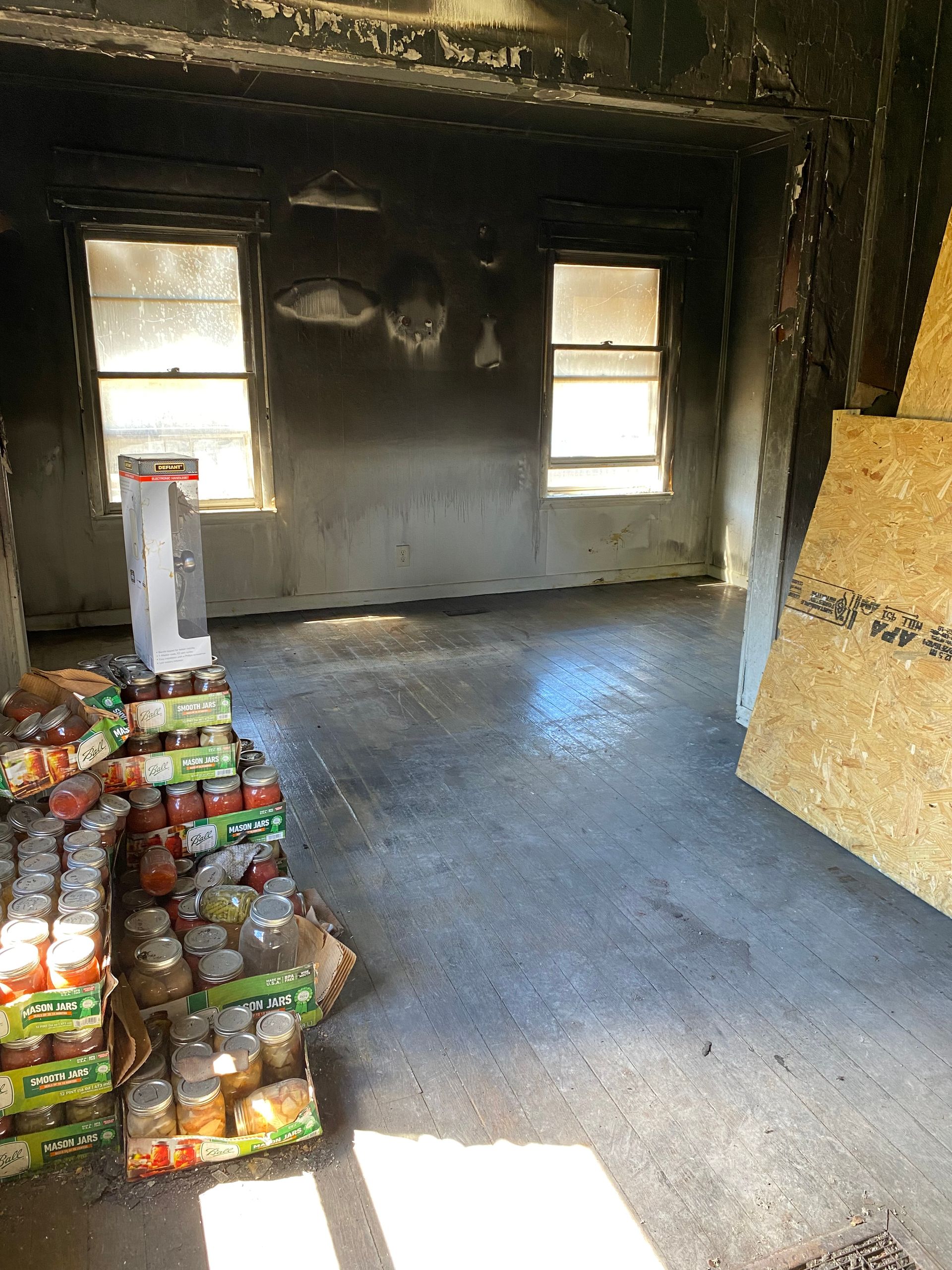 Empty charred room with two windows, concrete floor, and stacks of canned goods near the left wall