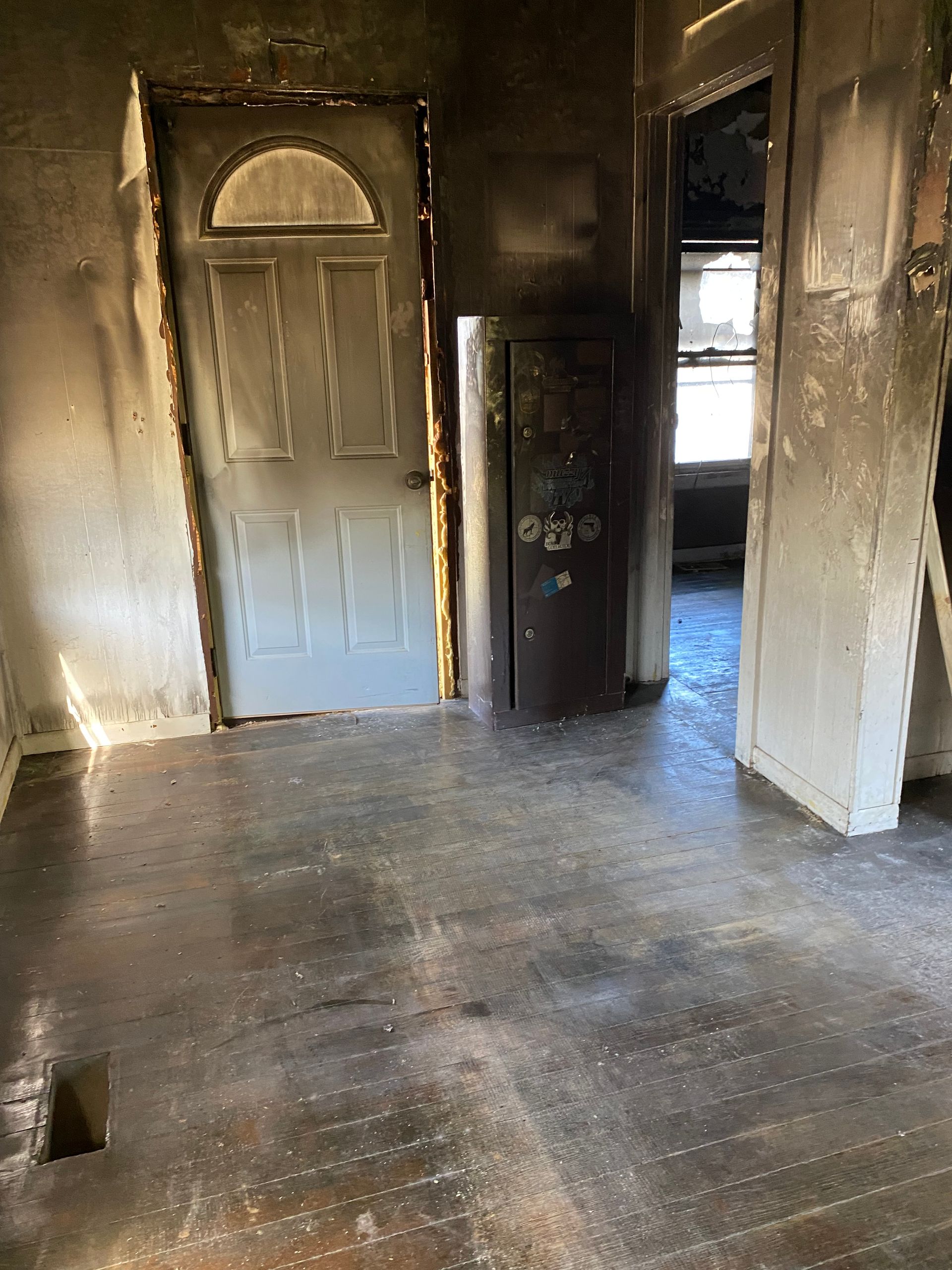 Burned empty room with soot-stained walls, a damaged door, and a dark cabinet near a doorway.