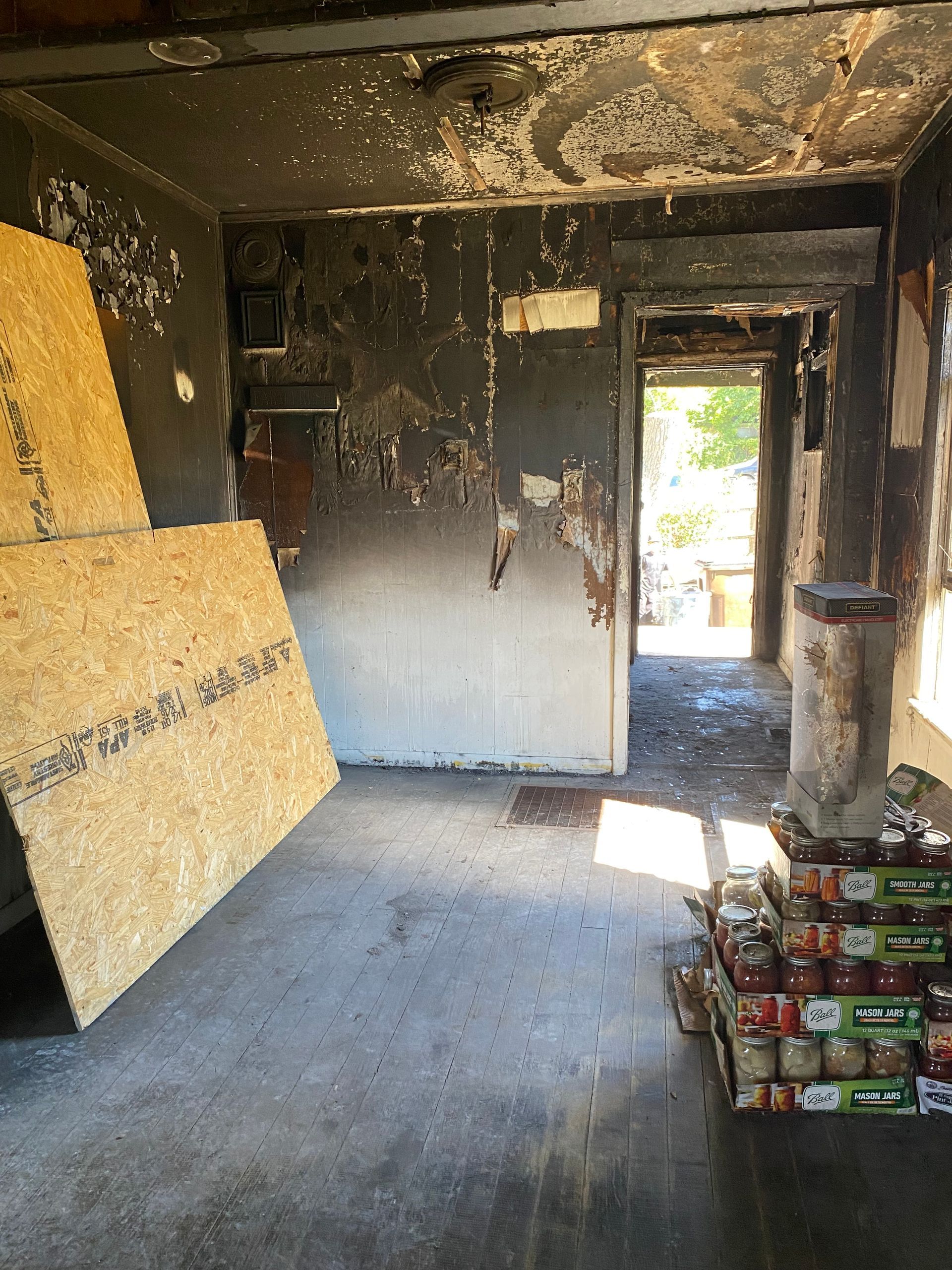 Charred, damaged room with burned walls, plywood leaning left, and stacked boxes near a doorway.