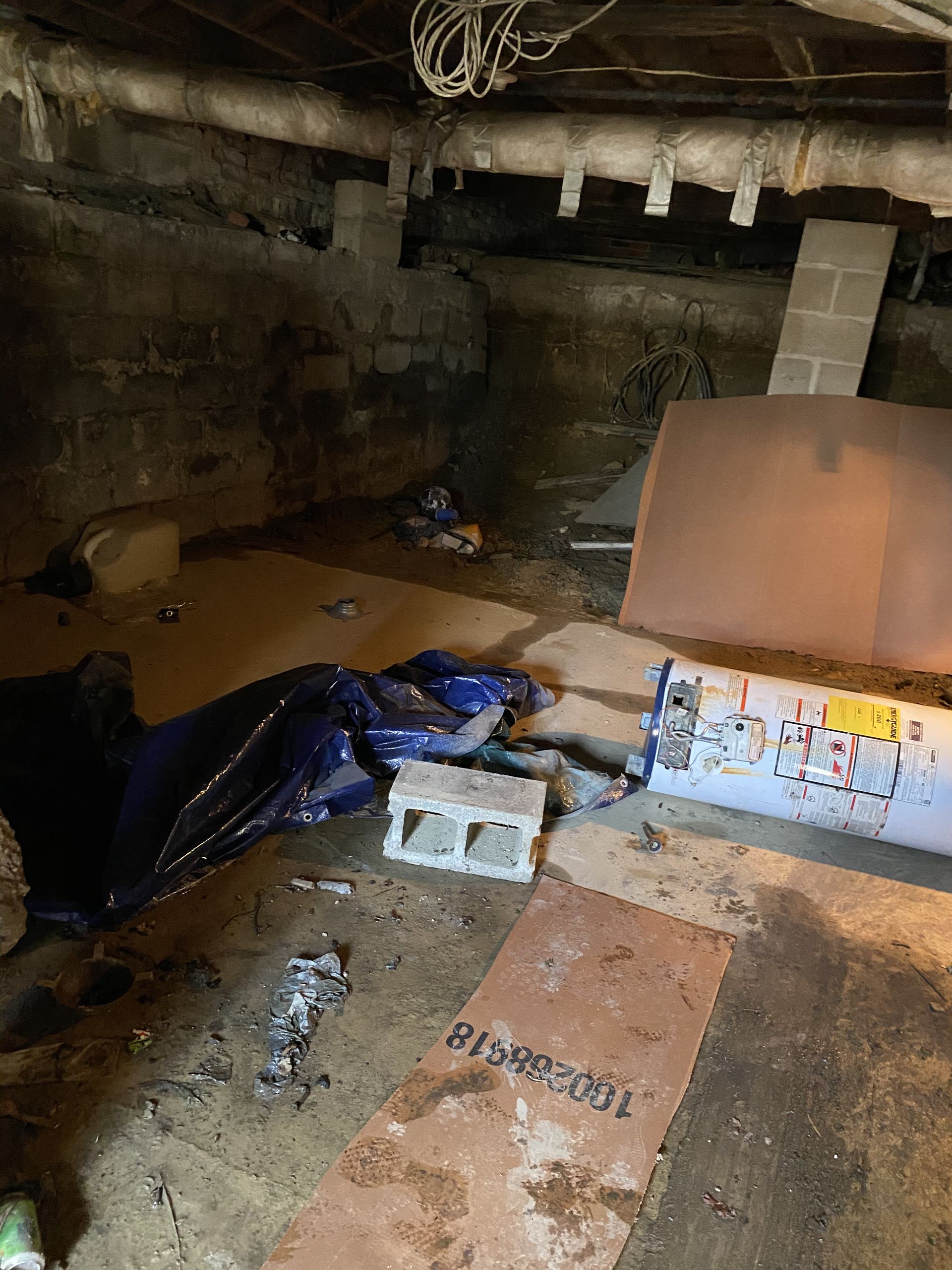 Dim basement cluttered with cardboard, debris, and a blue tarp under exposed pipes.
