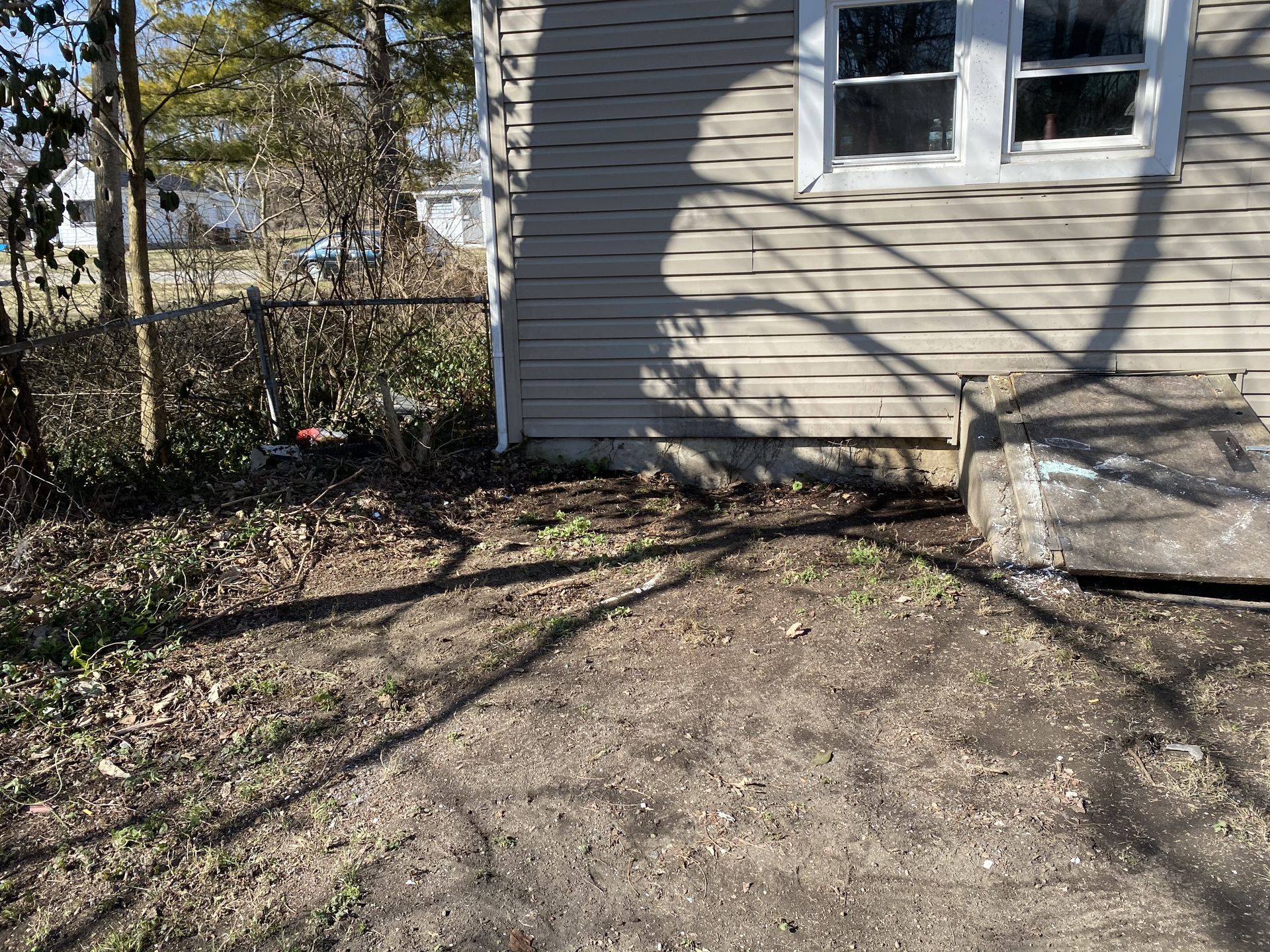 Side of a beige house with a white window, bare yard, and long tree shadows on the ground