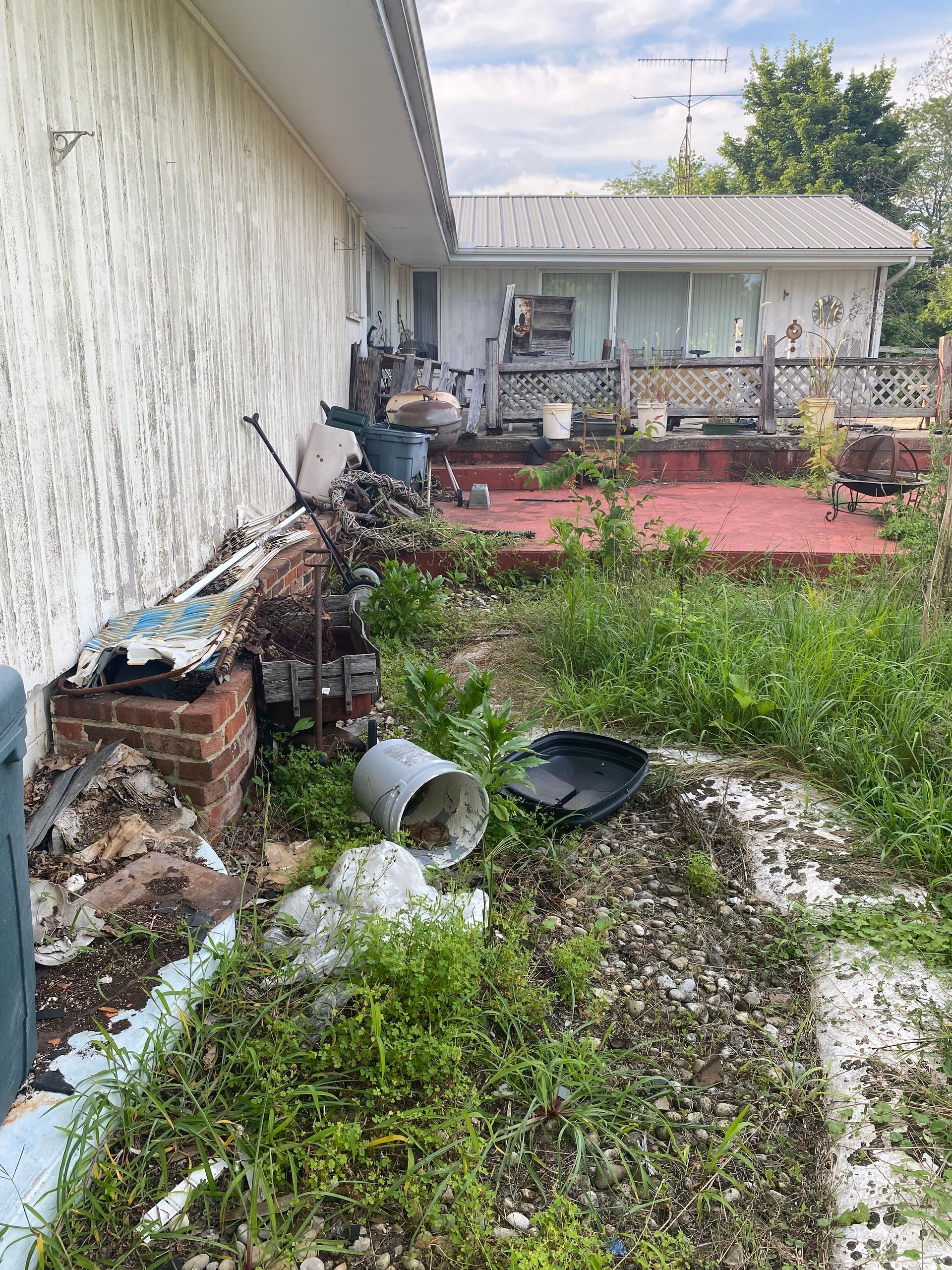 Damaged house exterior with broken wall, debris, and overgrown yard beside a red patio.