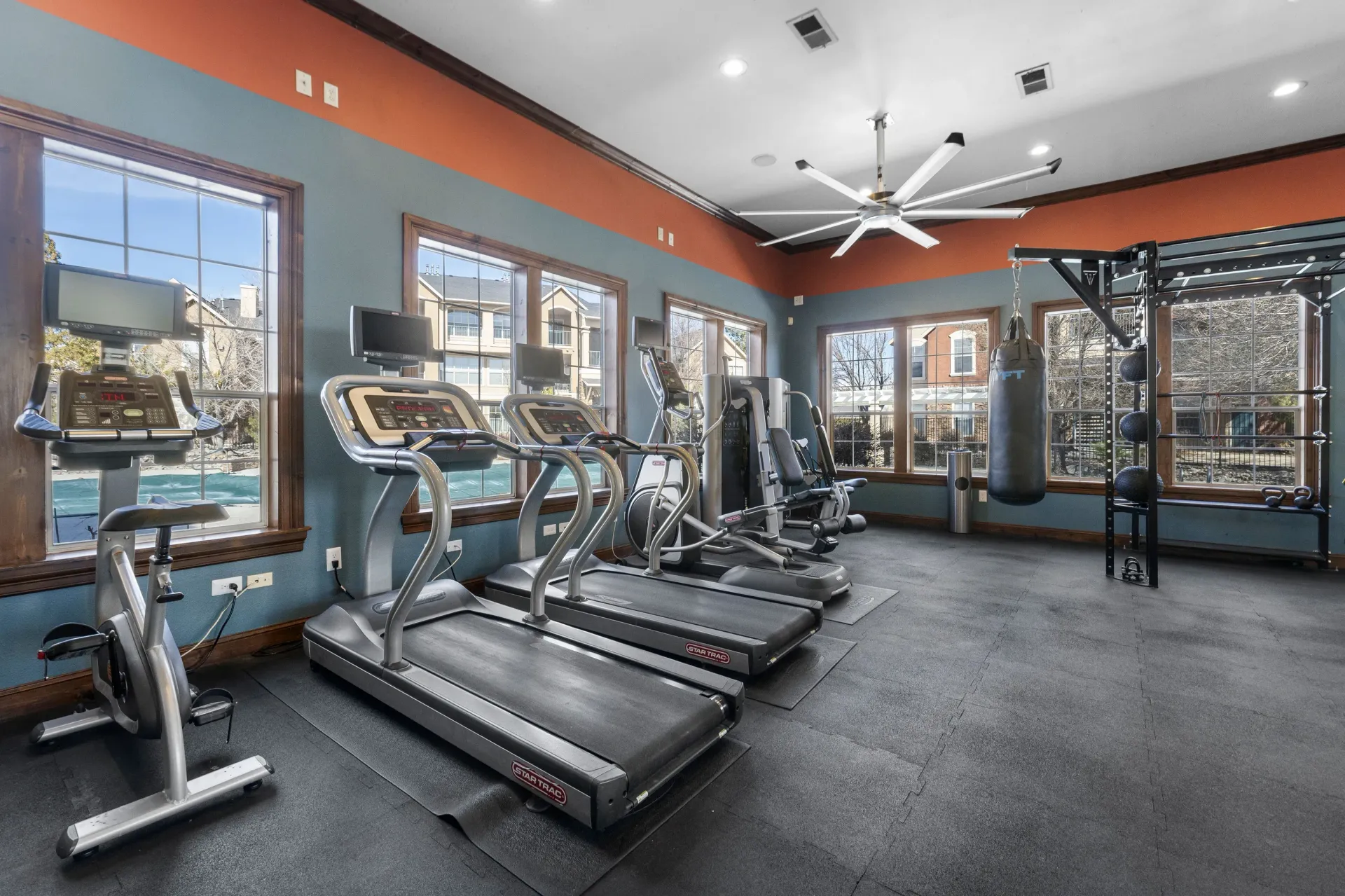 Fitness center with cardio machines, weight rack, and punching bag by windows.