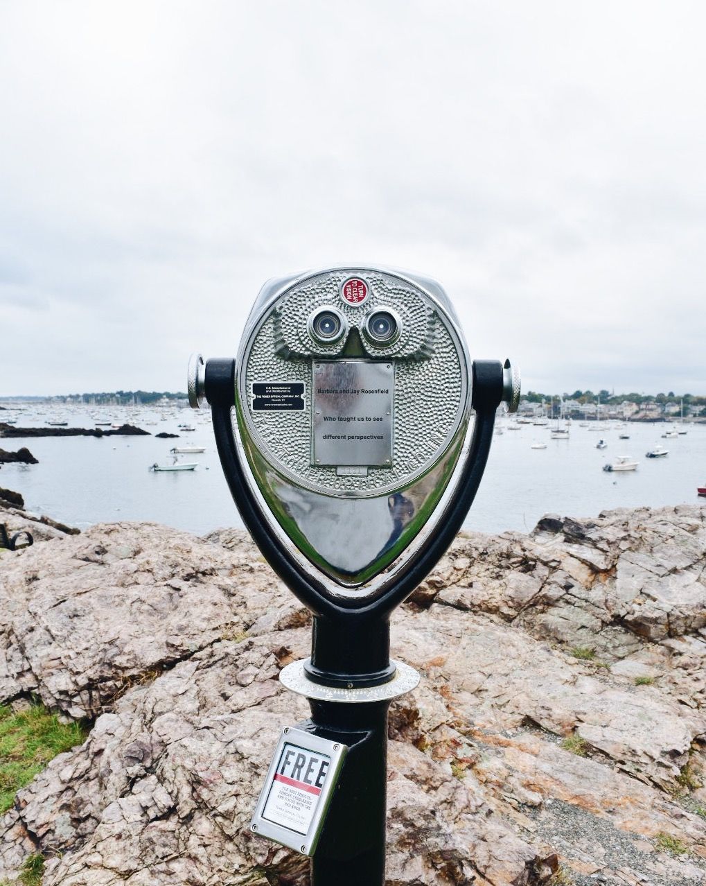 A metallic, coin-operated viewing scope stands on a rocky shore overlooking a harbor with distant boats under a grey sky.