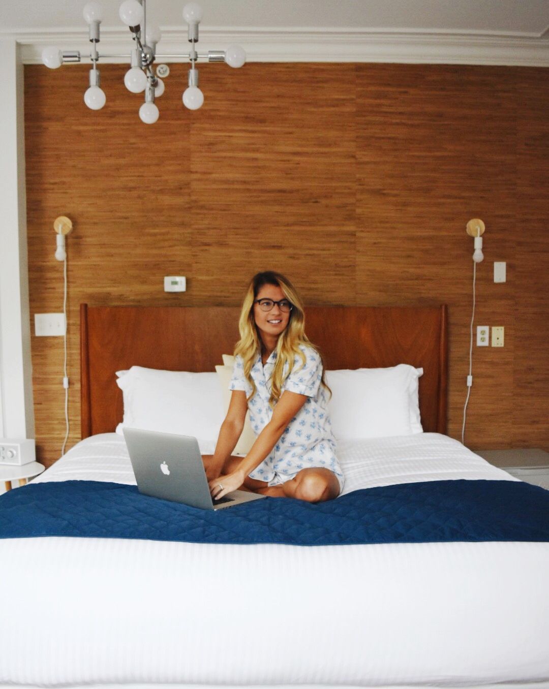 A smiling person wearing pajamas sits on a bed with a laptop, set against a wood-paneled wall with a modern chandelier.