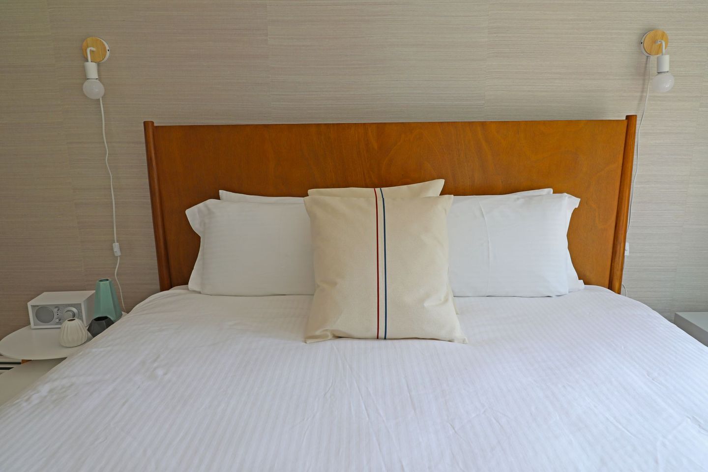 A bright bedroom featuring a queen bed with white linens and a blue runner, light wood furniture, and a television.
