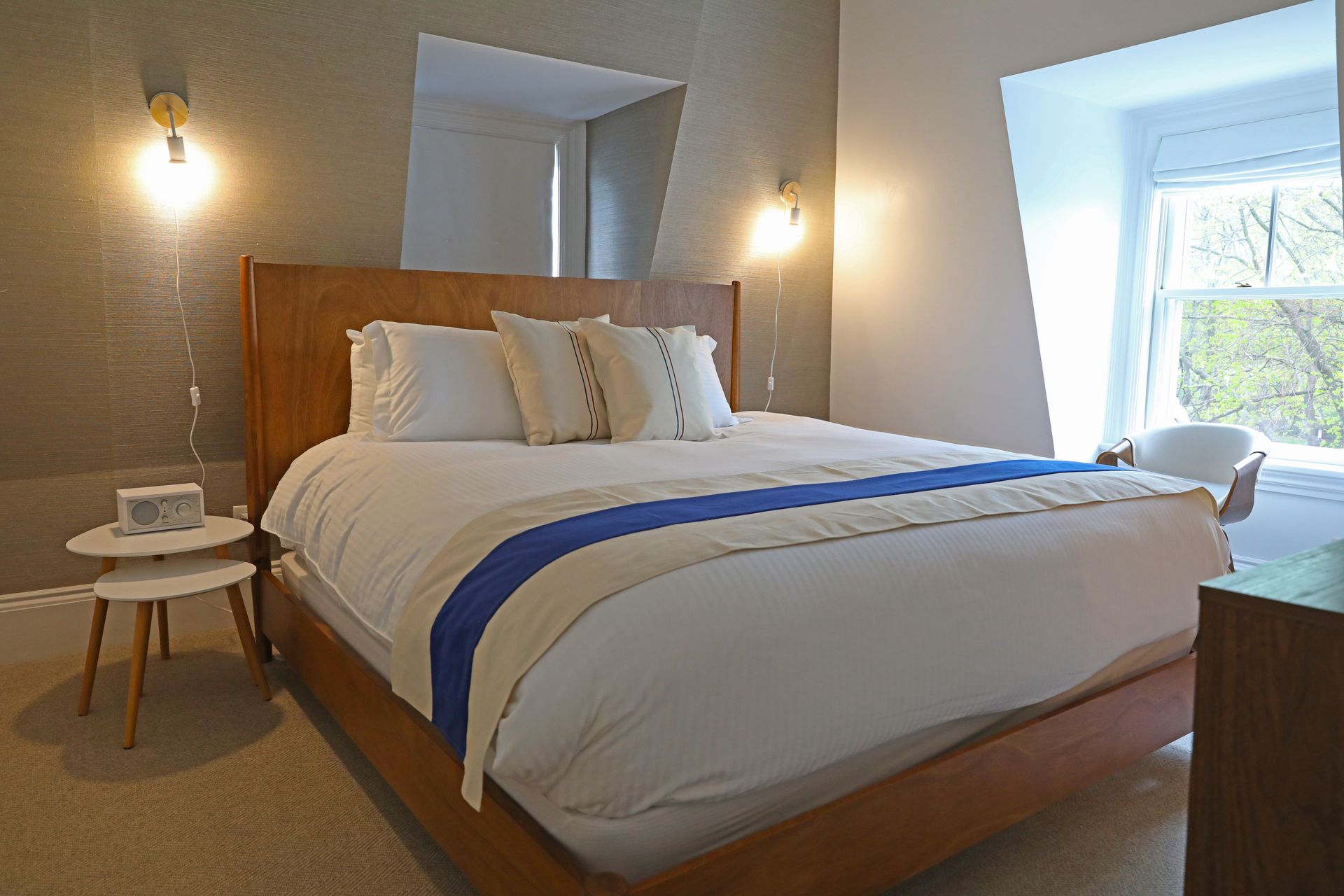 A bedroom with a wooden bed, white bedding with a blue stripe, two-tiered side table, and a window with natural light.