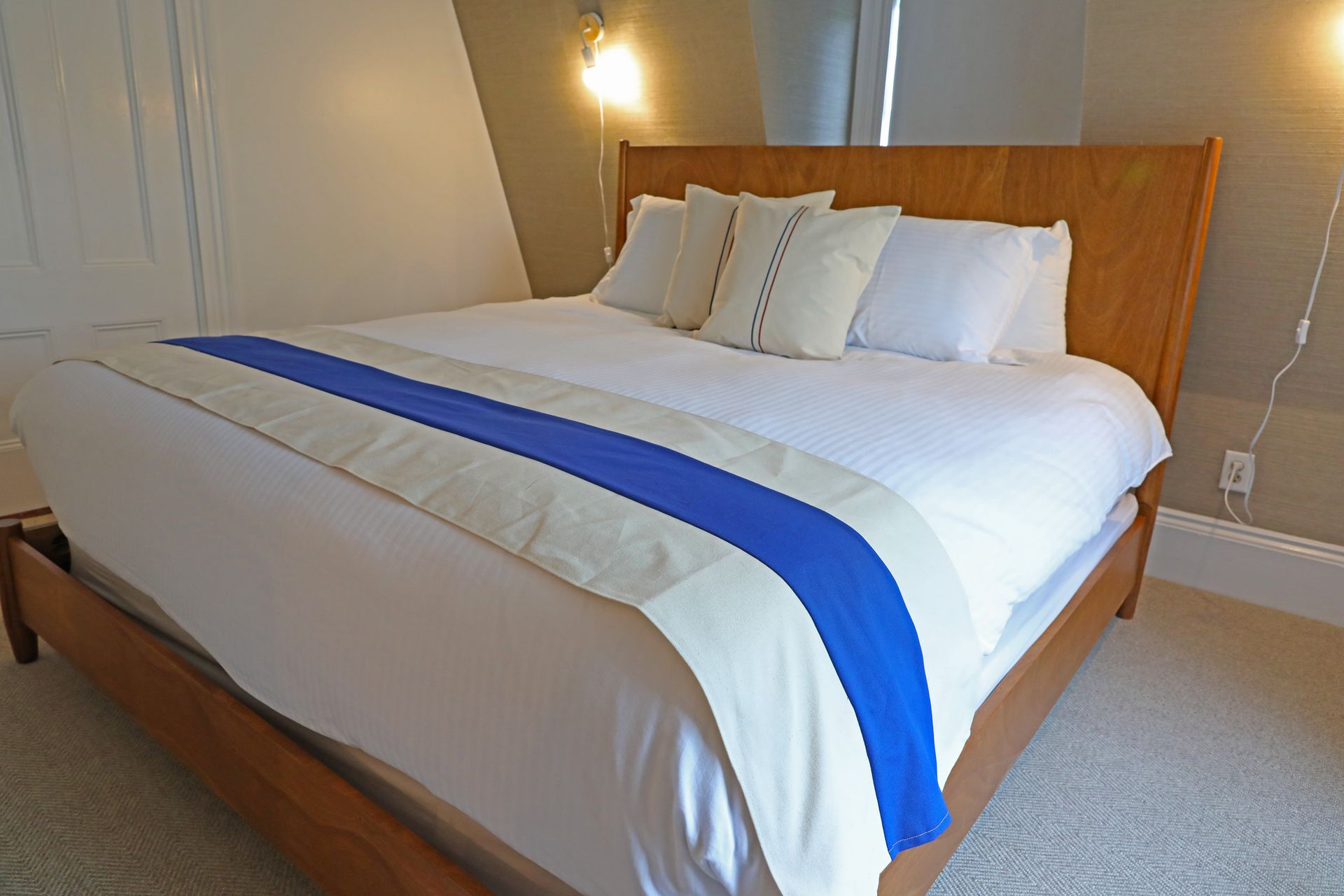 A modern bed with a wooden frame, white bedding, a blue and beige runner, and several decorative pillows in a bright room.