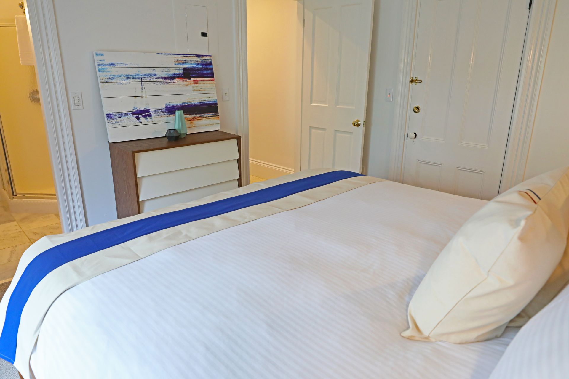 A white bed with a bright blue runner and a beige pillow faces a dresser topped with abstract art in a hotel room.
