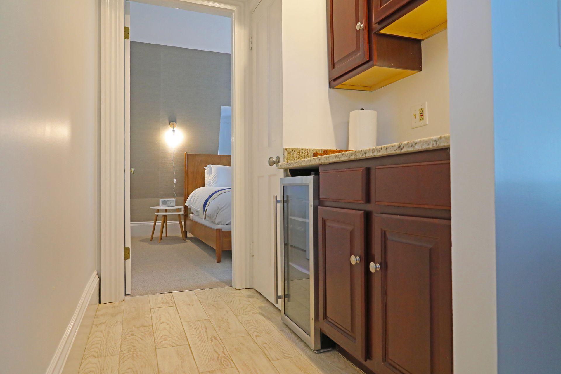 A hallway with light wood flooring leads to a bedroom, passing a small kitchenette with dark cabinets and a mini-fridge.