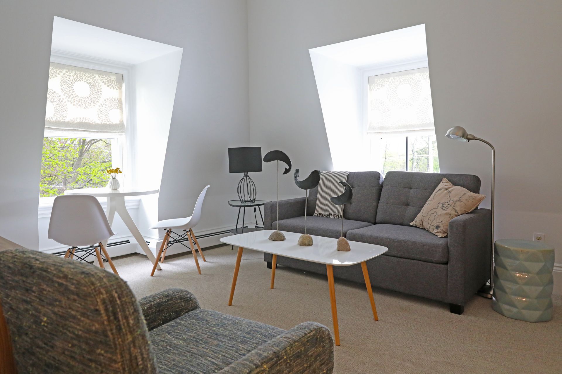 A bright living room with a grey sofa, coffee table, two white chairs by a window, and a textured armchair in the front.