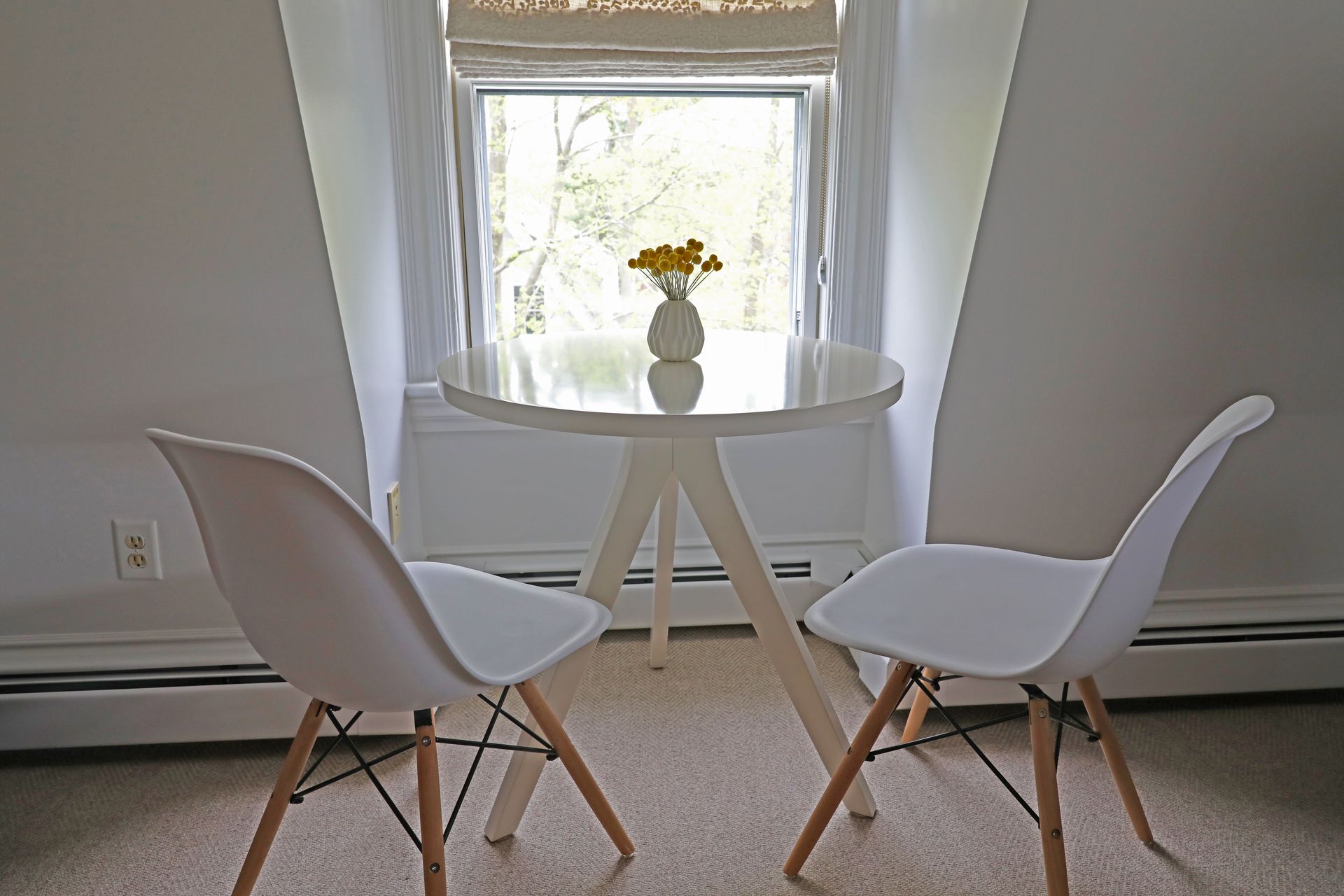 A small white round table and two white modern chairs sit in front of a window, with a small vase of yellow flowers.