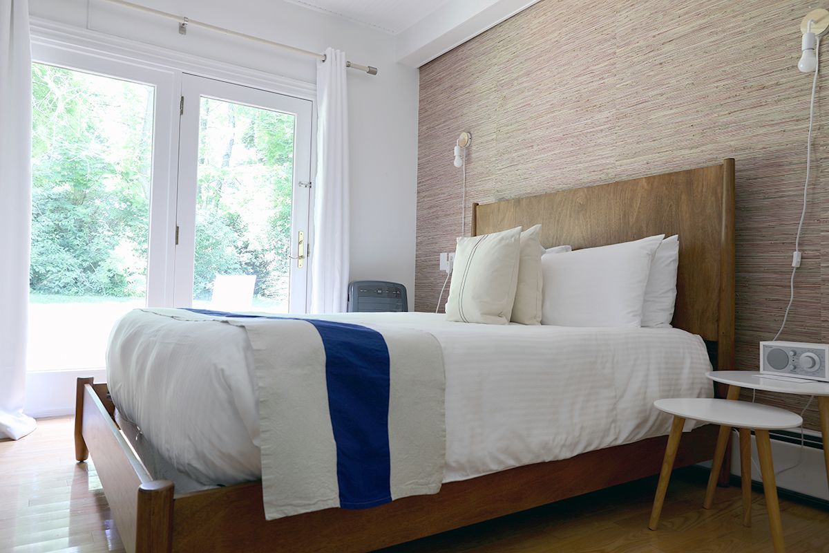 A bedroom with a wooden bed frame, white bedding with a blue runner, and a textured accent wall next to a glass door.