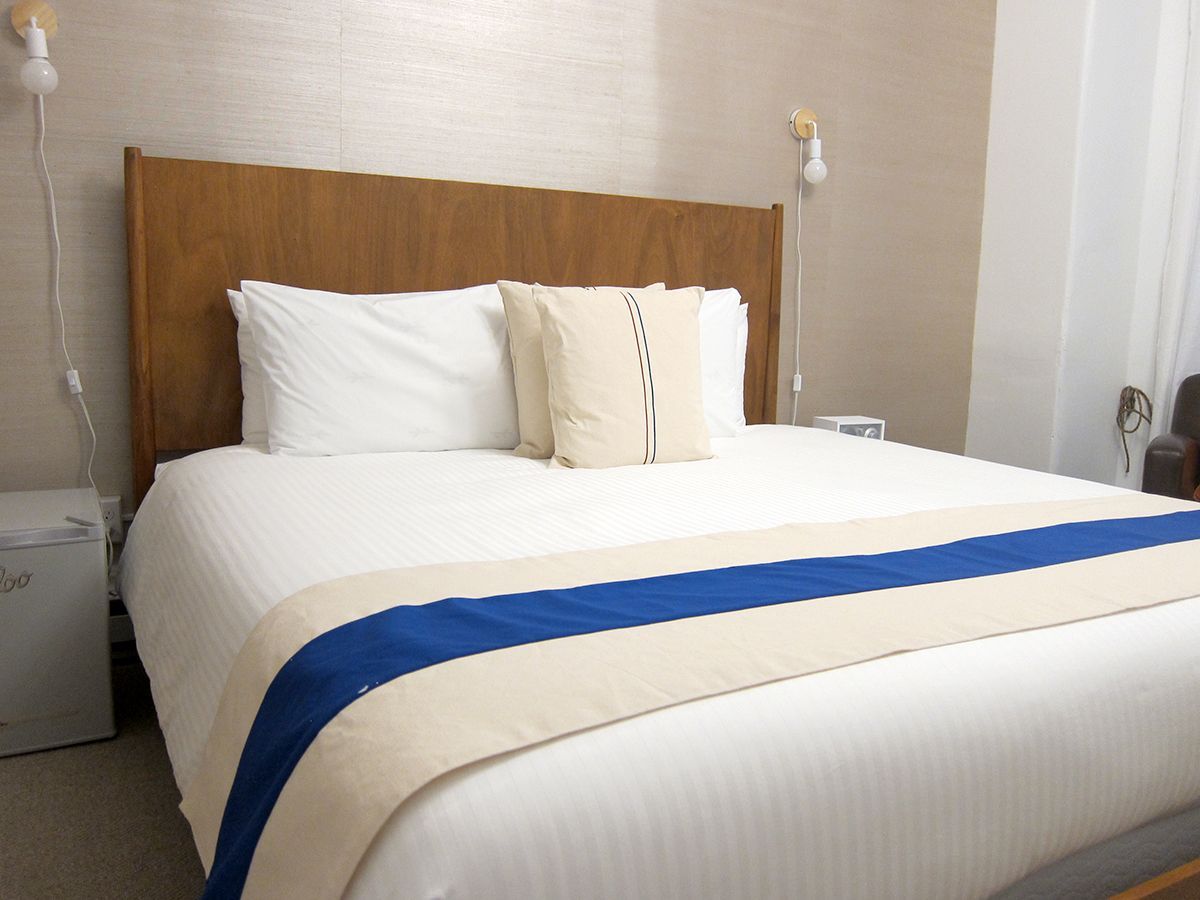 A hotel bed with a wooden headboard, white pillows, a cream accent pillow, and a beige runner with a blue stripe.