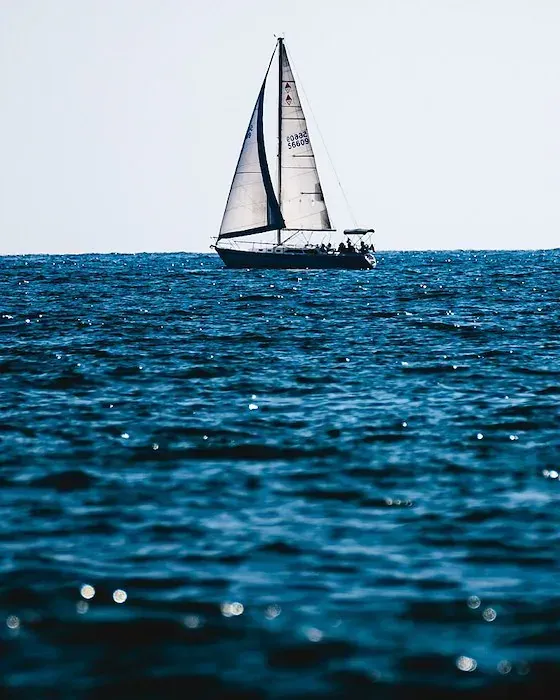 A lone sailboat with white sails floats on dark blue, rippled ocean waters under a clear sky.