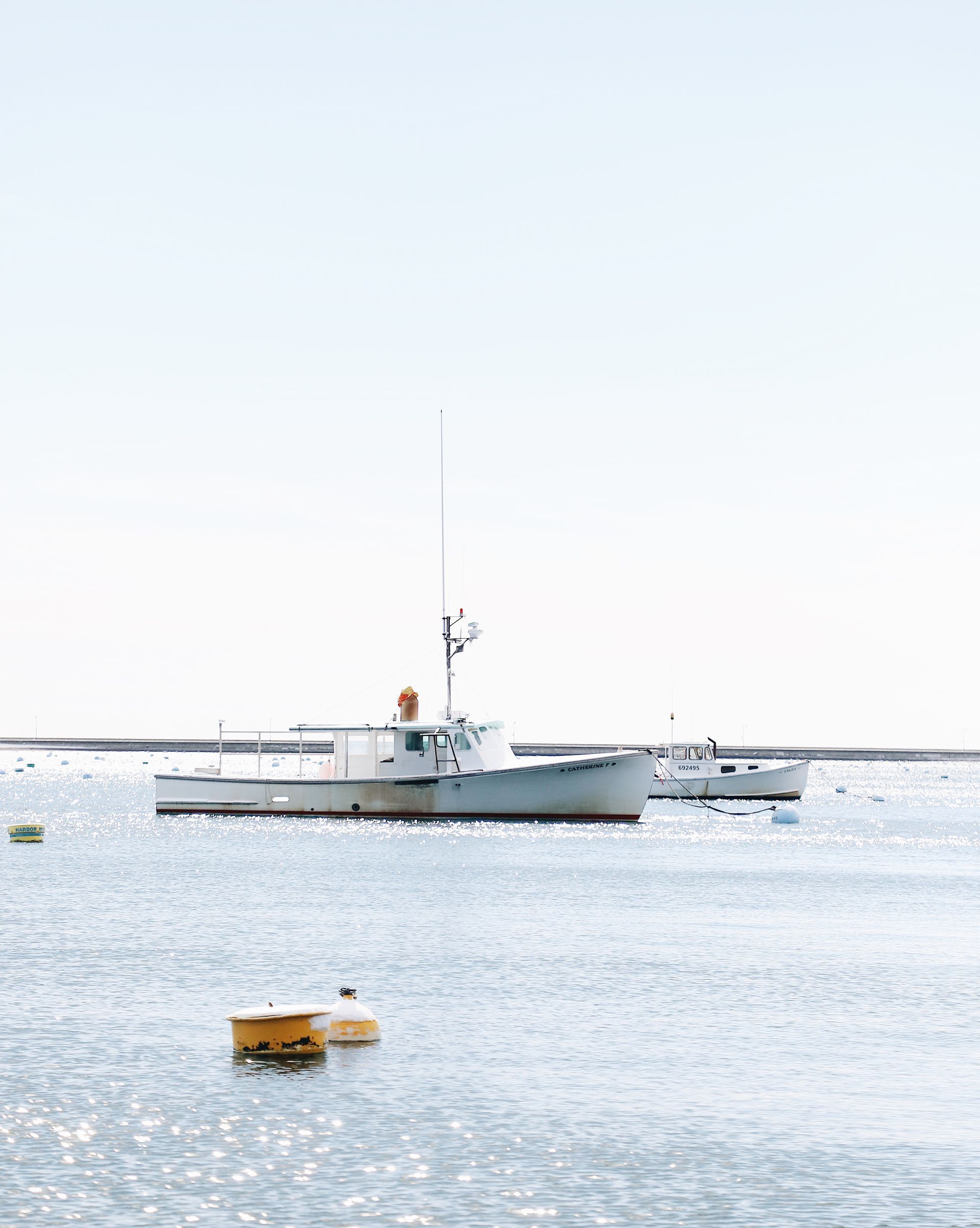 Two white motorboats anchored in a calm, sunlit harbor with a yellow mooring buoy floating in the foreground.