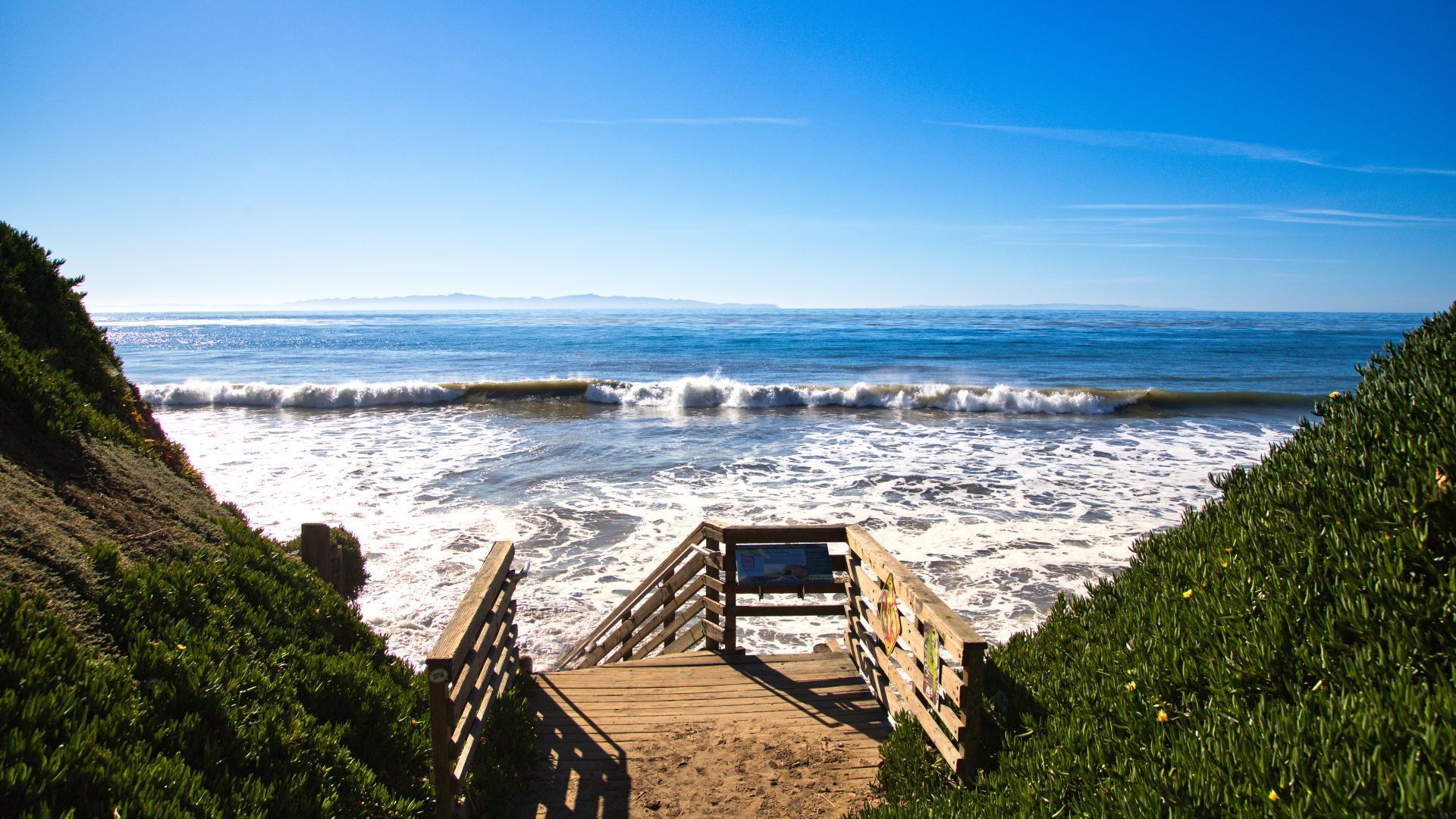 Wooden stairs leading to a sunny beach with waves and blue ocean under a clear sky