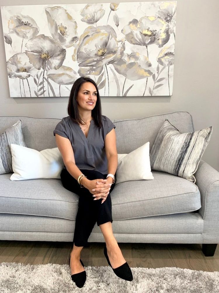 Woman seated on a gray sofa in a bright living room with floral wall art behind her.