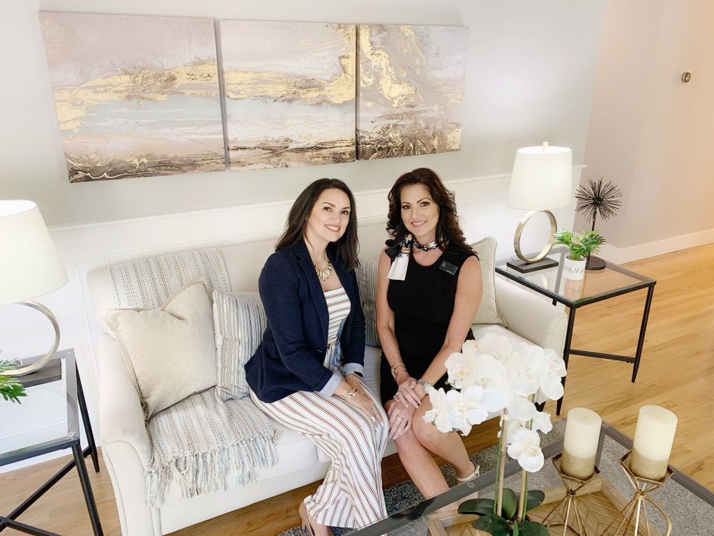 Two women sitting on a white sofa in a bright living room with gold and white decor.