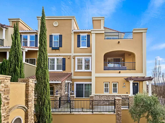 Row of tan stucco townhouses with balconies, front gate, and tall cypress trees under a blue sky