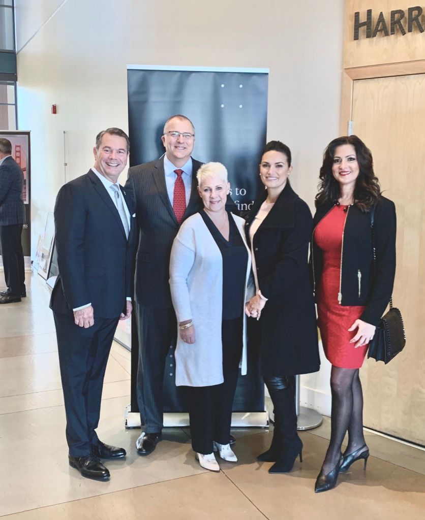 Five people in business attire posing in a lobby, standing in front of a gray backdrop.