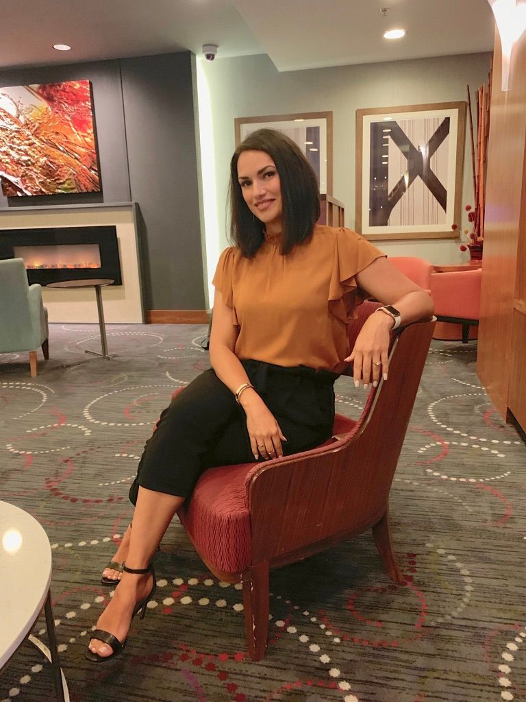 Woman seated in a red chair in a modern lobby, wearing an orange top and black pants, with artwork in the background.