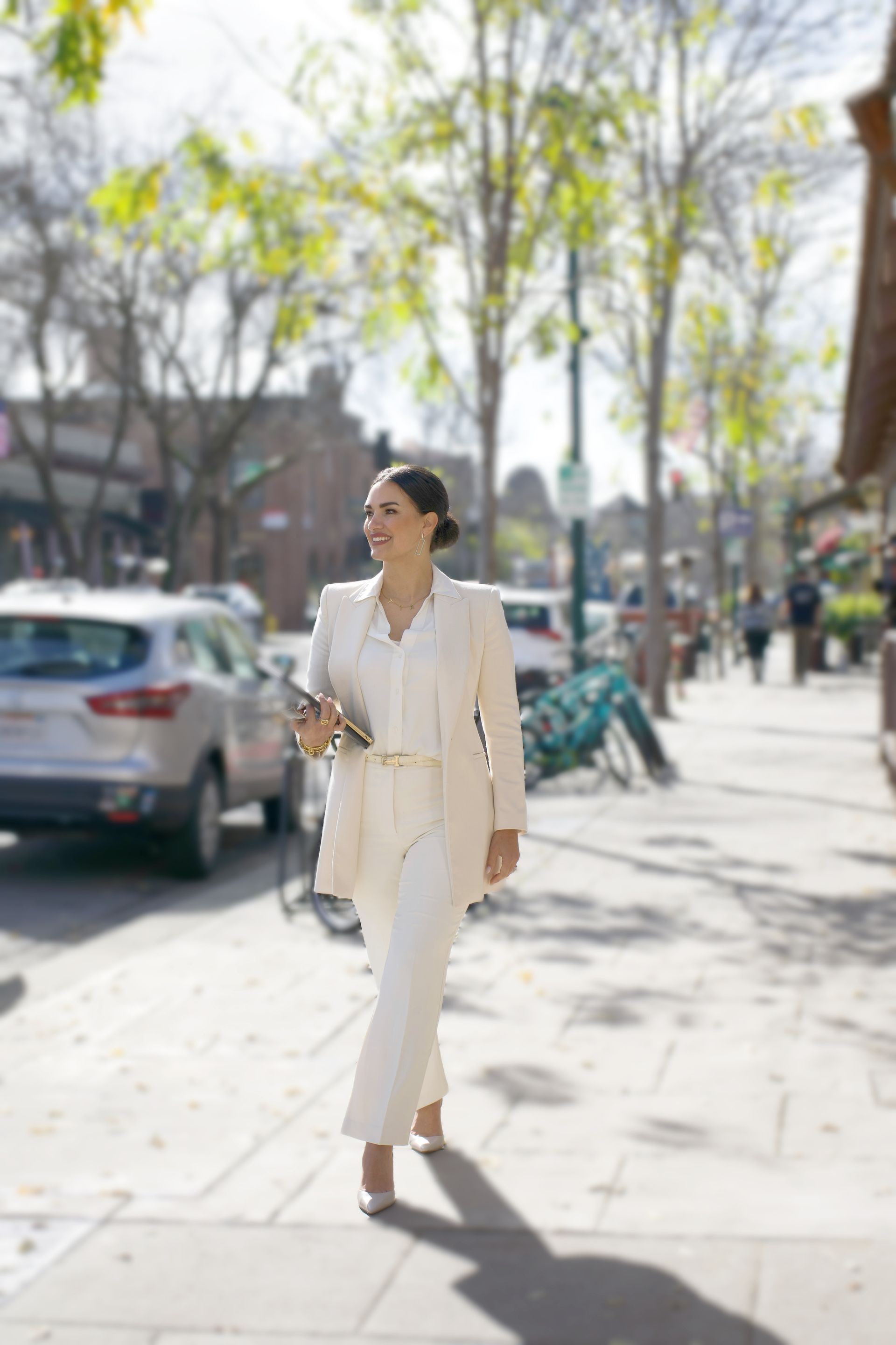 Person in a white suit walking on a sunny city sidewalk with trees and parked cars nearby