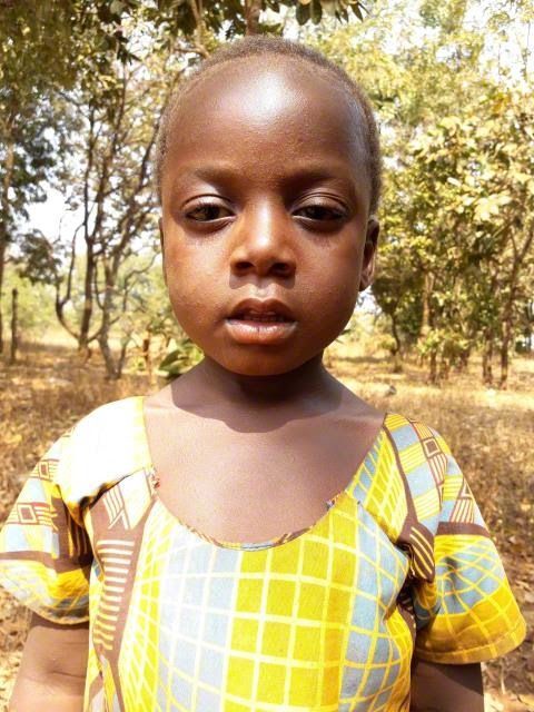 Child standing outdoors in a yellow patterned shirt, looking at the camera, with trees in the background