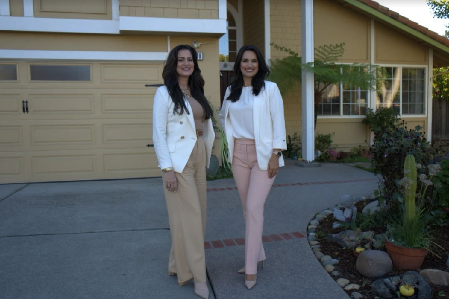 Two women in white blazers and light pants standing outside a house by the driveway