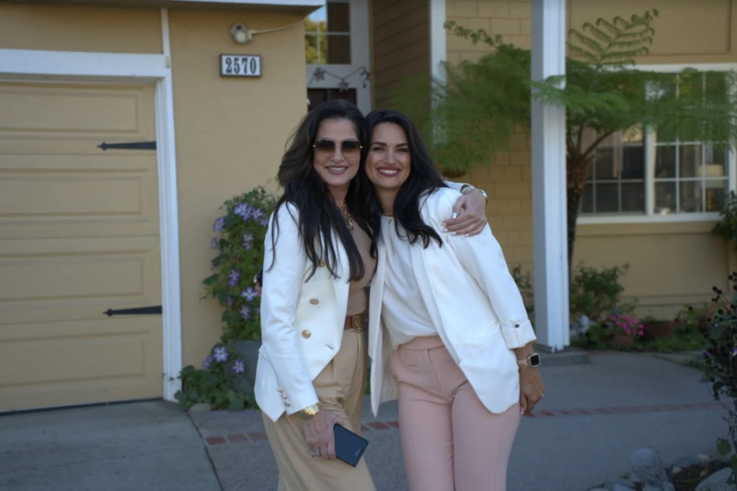 Two women in white jackets smiling and posing outside a house, one with an arm around the other.