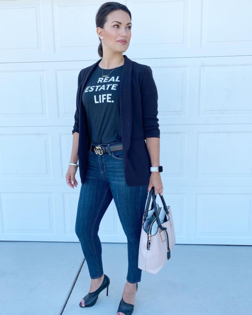 Woman standing outdoors in black jacket, graphic tee, jeans, and heels, holding a handbag.