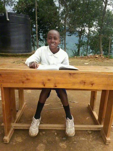 Child smiling behind a wooden desk outdoors, with open books and trees in the background