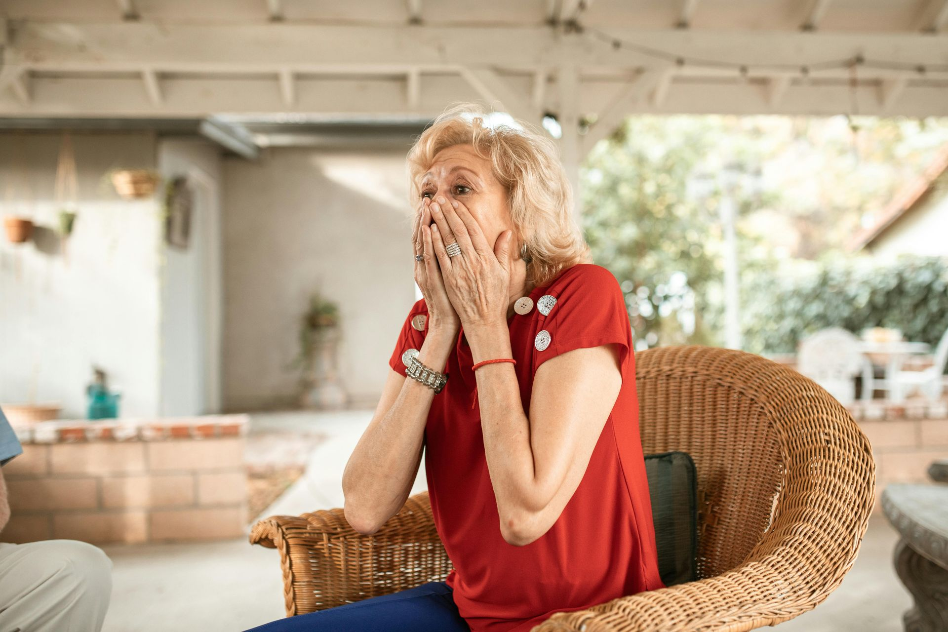 Woman with surprised expression covers mouth; seated outdoors in a wicker chair.