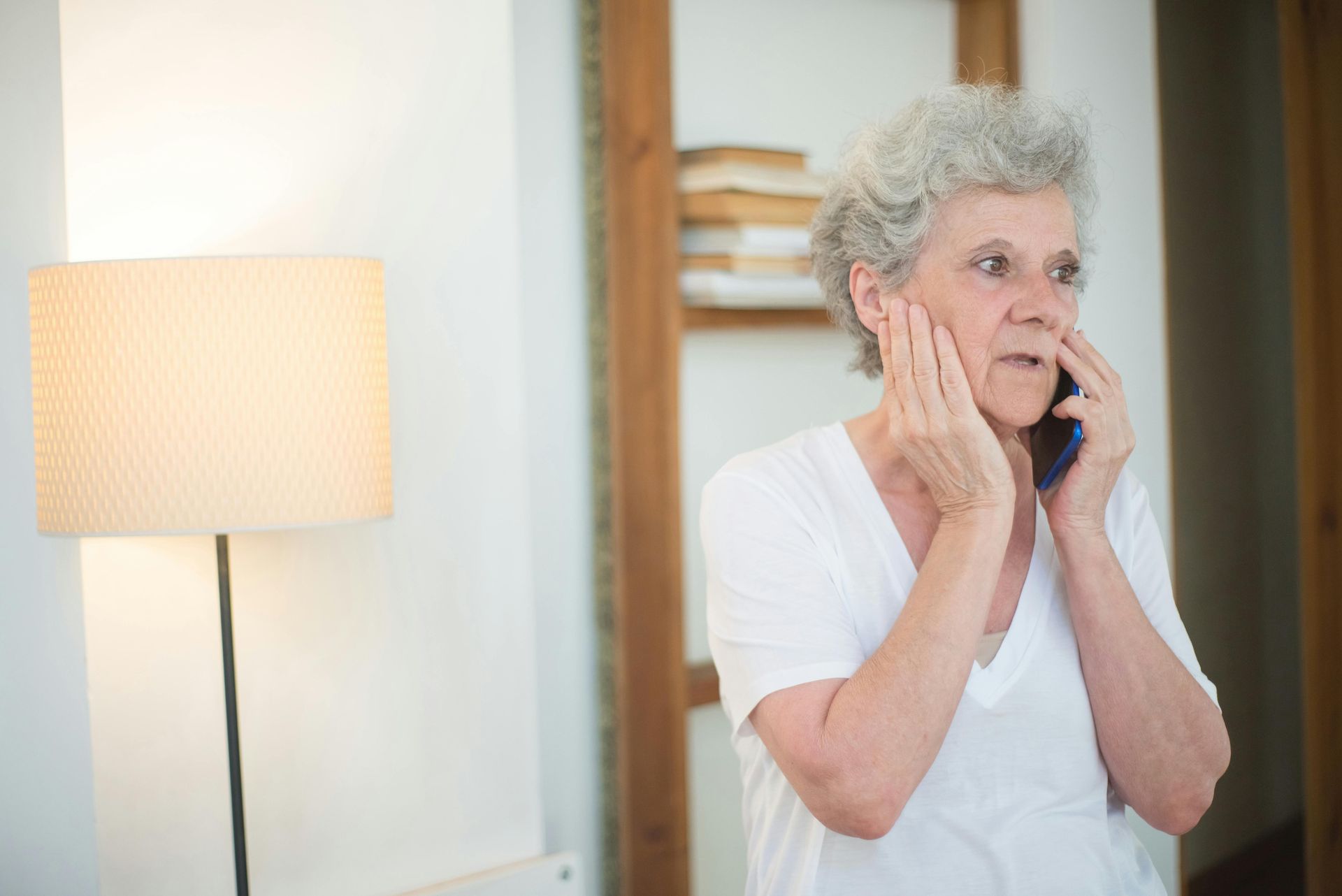 Woman in white shirt talking on phone, touching her face, looking concerned.