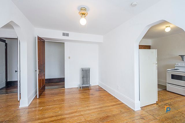 An empty living room with hardwood floors and white walls leading to a kitchen.