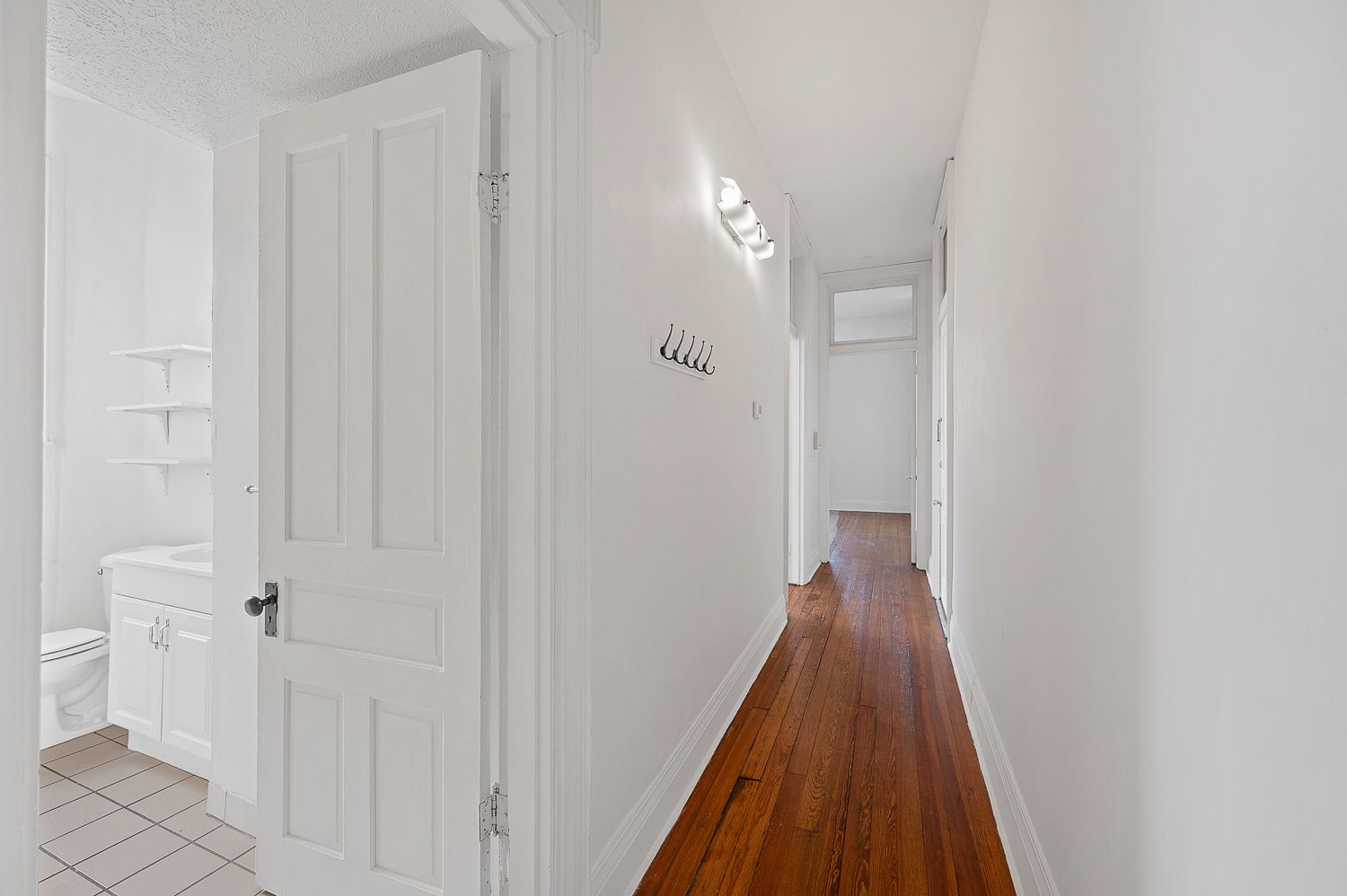 A long hallway with white walls and wooden floors leading to a bathroom.