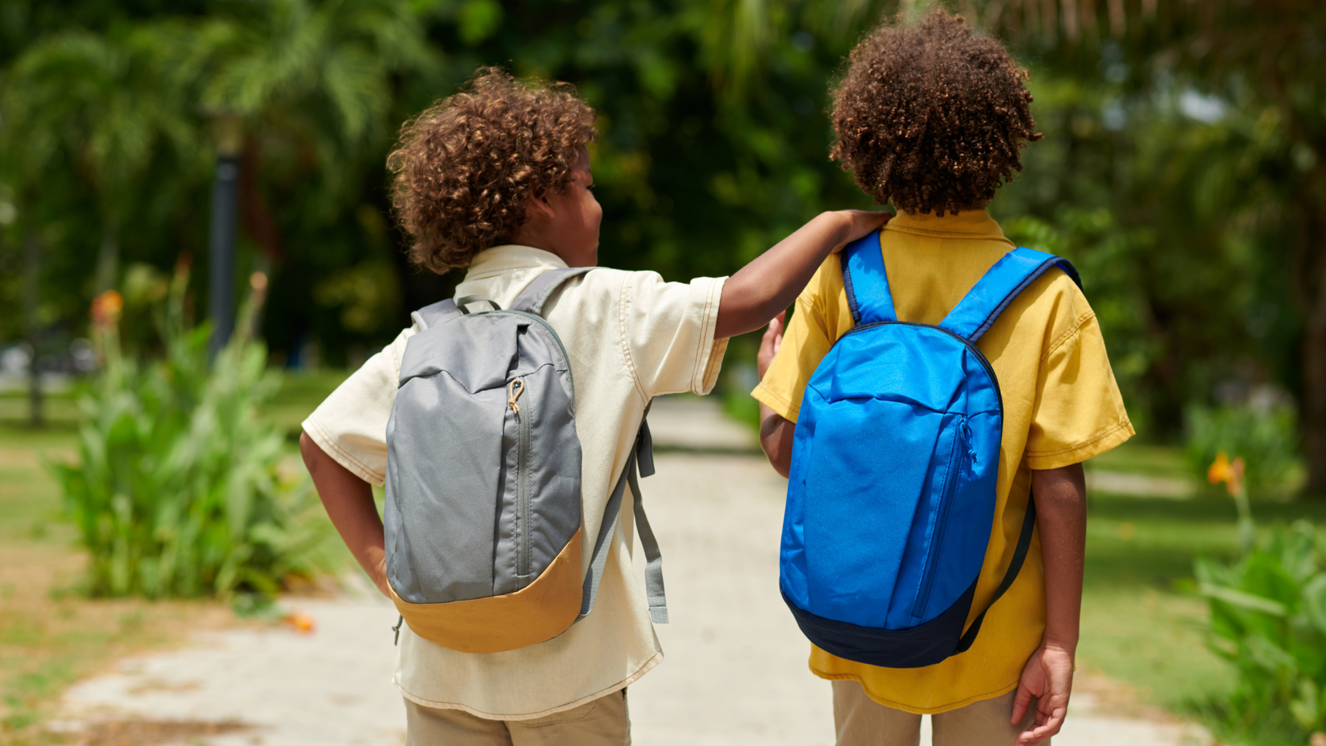 Two children with curly hair walk down a path in a park, one wearing a gray backpack and the other a blue backpack.