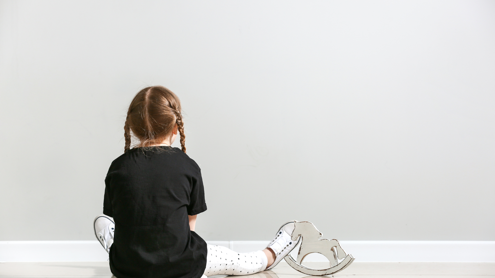 A person with braided hair sits on a floor in front of a white wall next to a small wooden rocking horse.