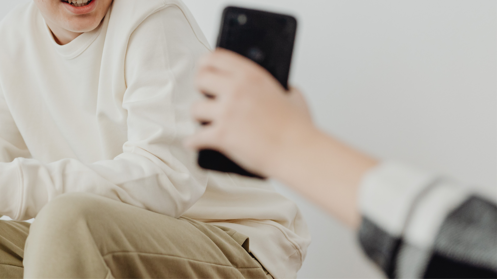 A close-up of a hand holding a black smartphone up toward a person wearing a beige sweater.