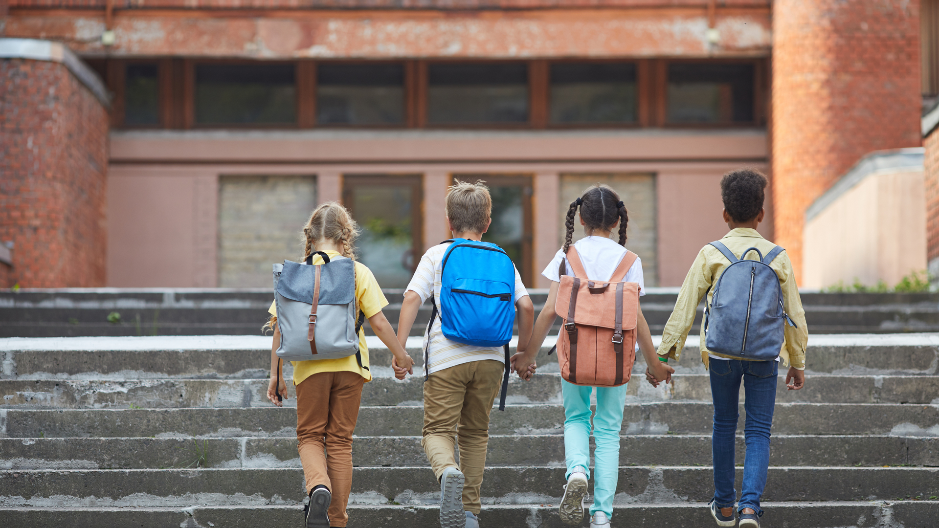 Four students with backpacks walking up steps toward a brick campus building