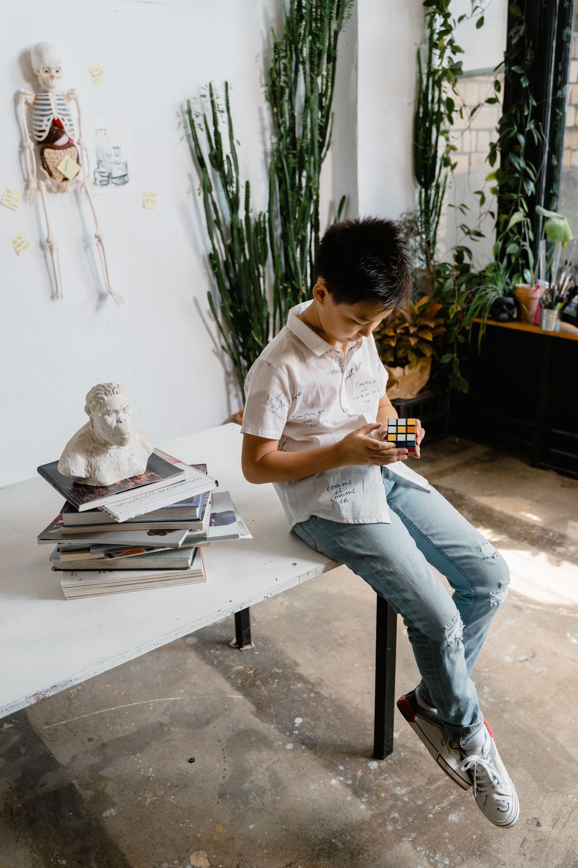 Person in a white shirt and jeans sits on a bench, looking at a phone in a bright plant-filled room.