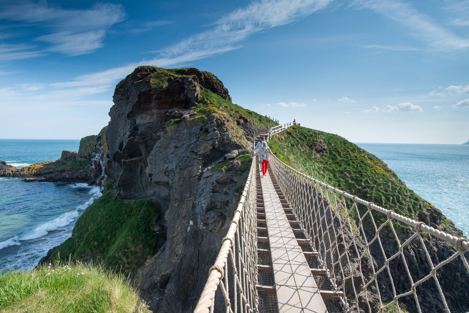A person walks across a rope bridge to a rocky island on a sunny day; ocean on either side.