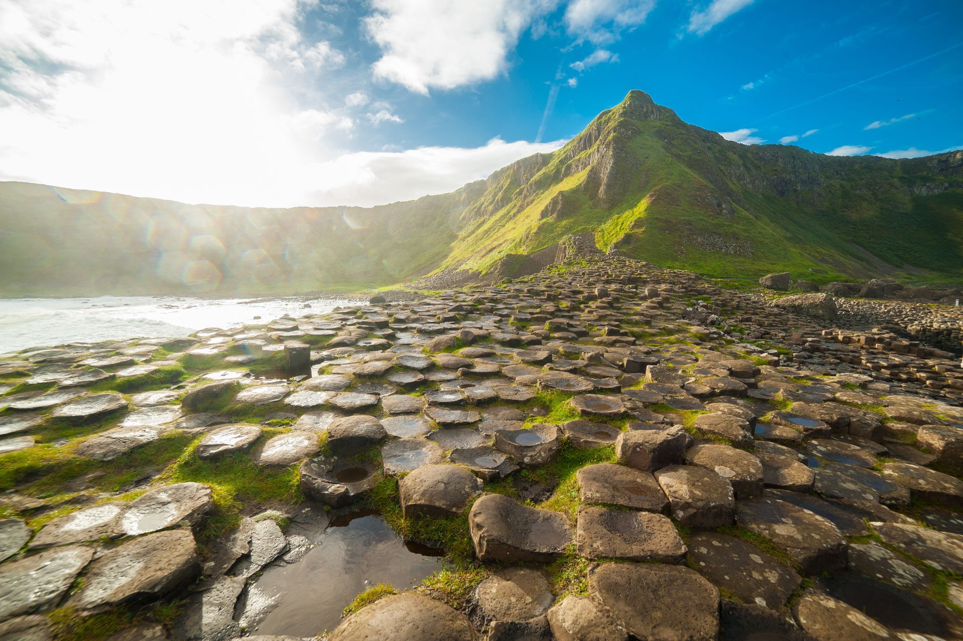 A rocky shoreline of hexagonal basalt columns leads to a green mountain, under a bright blue sky.