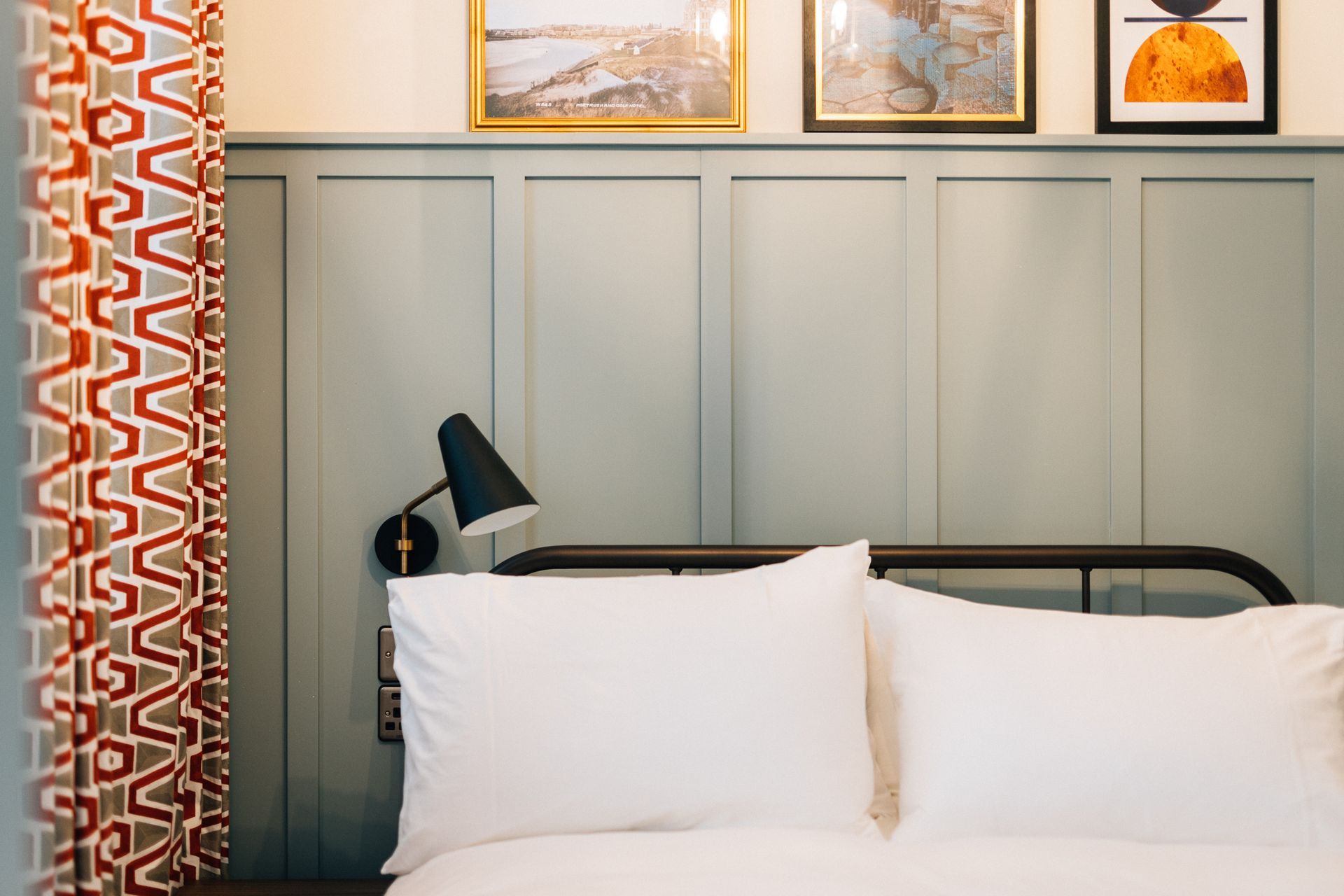Bedroom with a bed, two white pillows, and a black lamp. Light blue wall paneling, artwork, and patterned red curtains.
