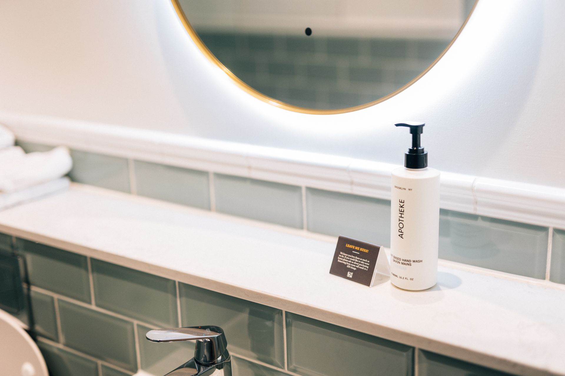 Bathroom counter with a circular mirror, a bottle of lotion, and a card.